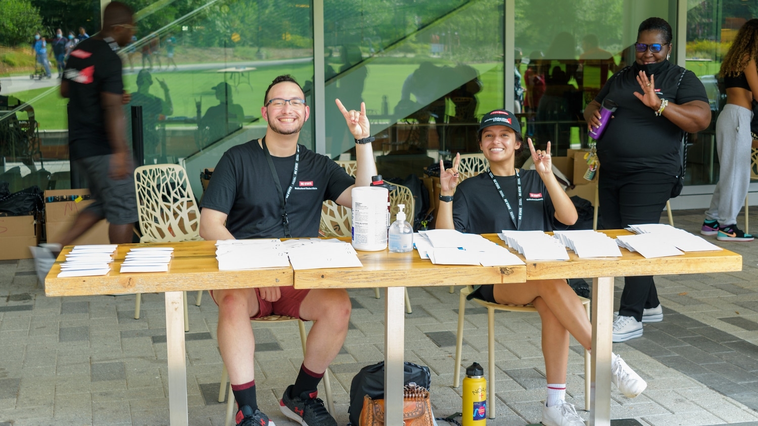 Three people wearing Symposium t-shirts smile at the camera and throw up wolfies while they wait to welcome incoming students.
