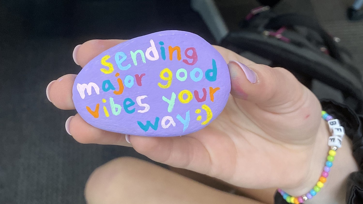 A student holds a painted purple rock with the message "sending major good vibes your way :)" on it. Each letter is in a different color. This photo is a close-up of the rock and the student's hand.